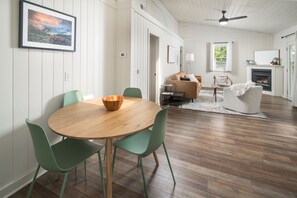 A dining area with a round wooden table and green chairs, a sofa, and a fireplace in a room with wooden floors and white walls. - The Victorian Inn Blowing Rock (Blowing Rock)