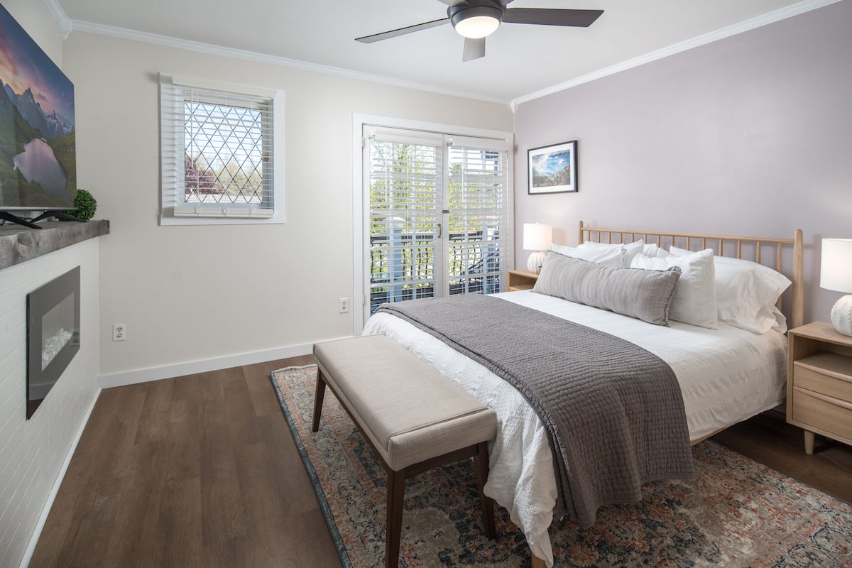A bedroom with a bed, bench, bedside tables, a ceiling fan, a window with blinds, and a door to an outdoor area.