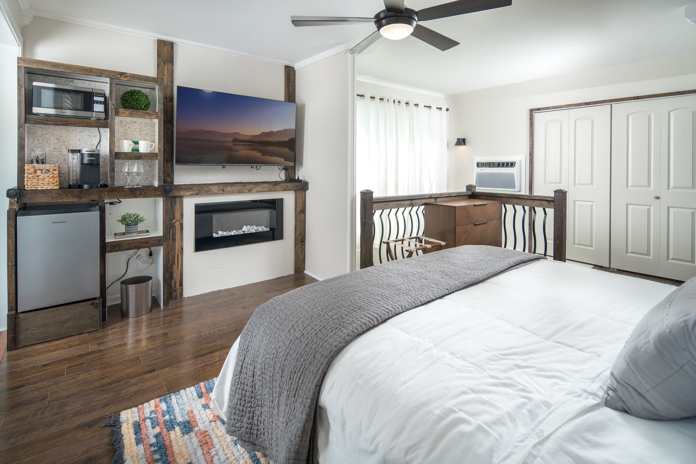 A bedroom with a bed, a TV, a ceiling fan, a fireplace, a kitchen area with a microwave, and a refrigerator.
