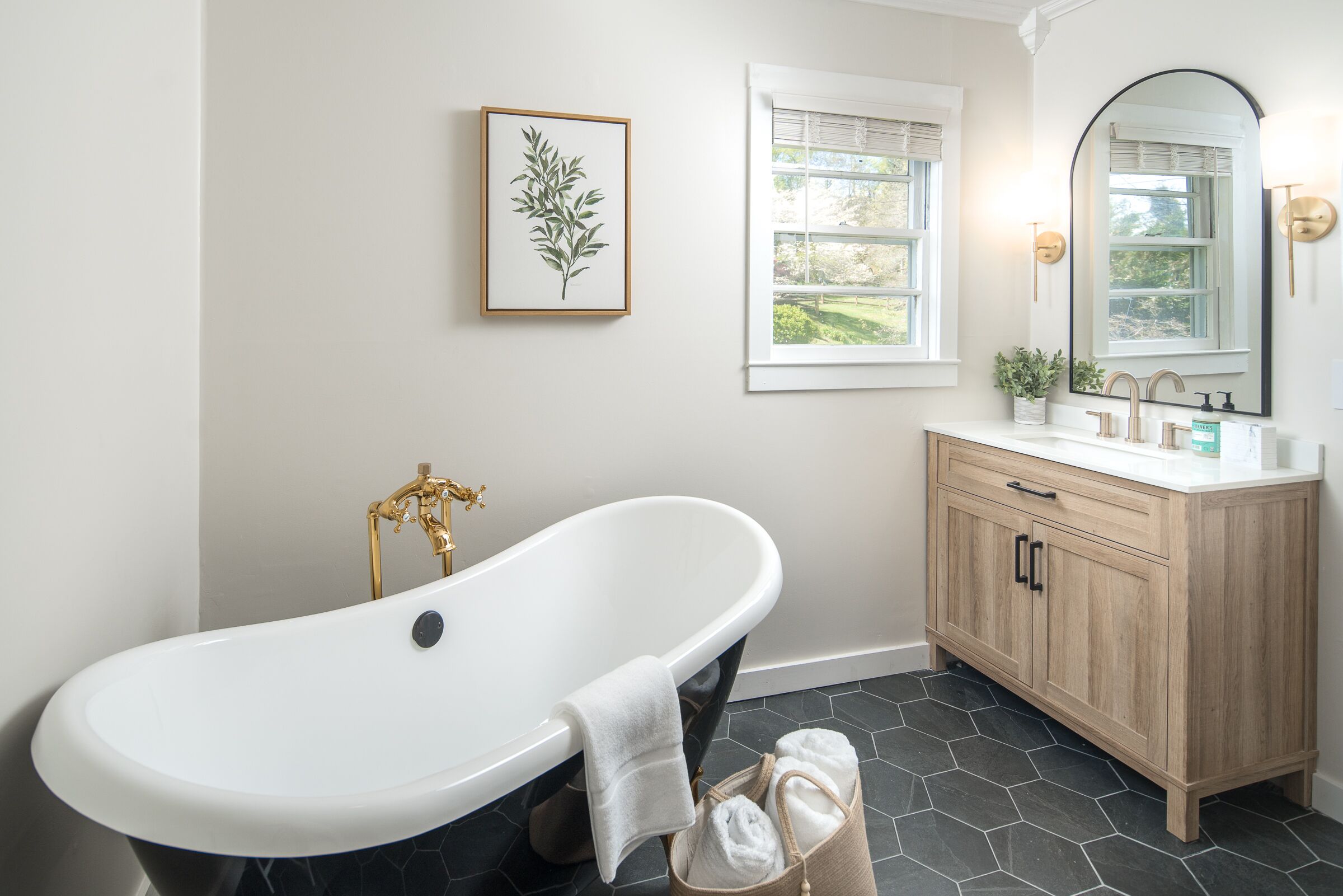 Ein Badezimmer mit freistehender Badewanne, einem Holzwaschtisch, einem gerahmten Pflanzenprint an der Wand und einem Fenster mit Jalousien.
