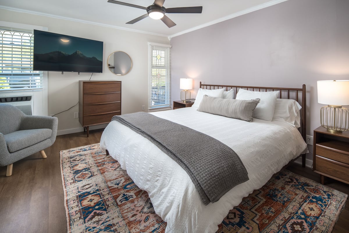 A bedroom with a bed, a television, a ceiling fan, a dresser, a chair, and a window with blinds.