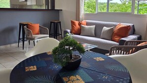 Ein modernes Wohnzimmer mit Sofa, Sesseln, einem Esstisch mit Blumentopf und einer Küche im Hintergrund.