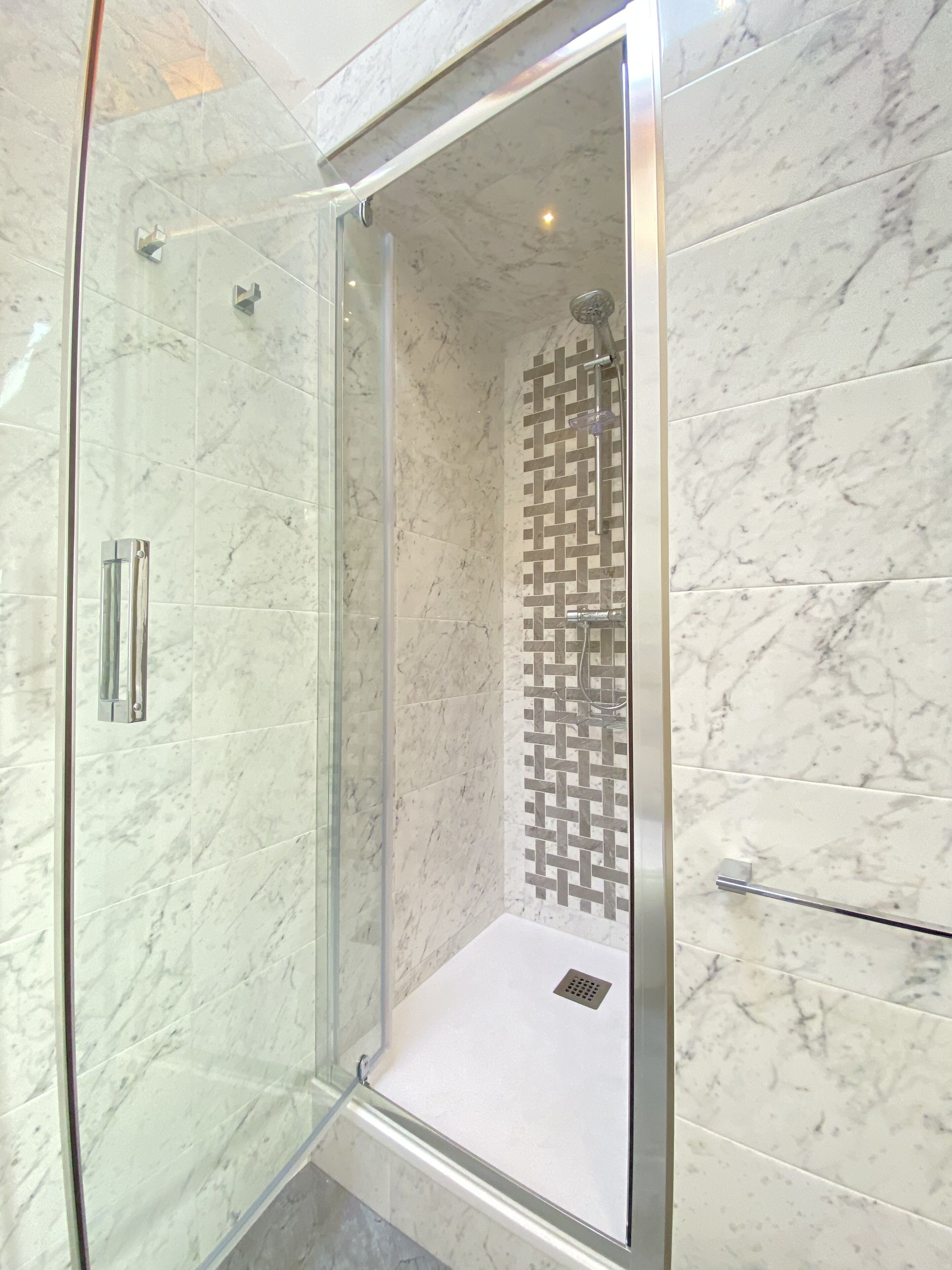 Une douche fermée par des parois de verre, avec un mur recouvert de carreaux de marbre et une pomme de douche en forme de grille métallique.