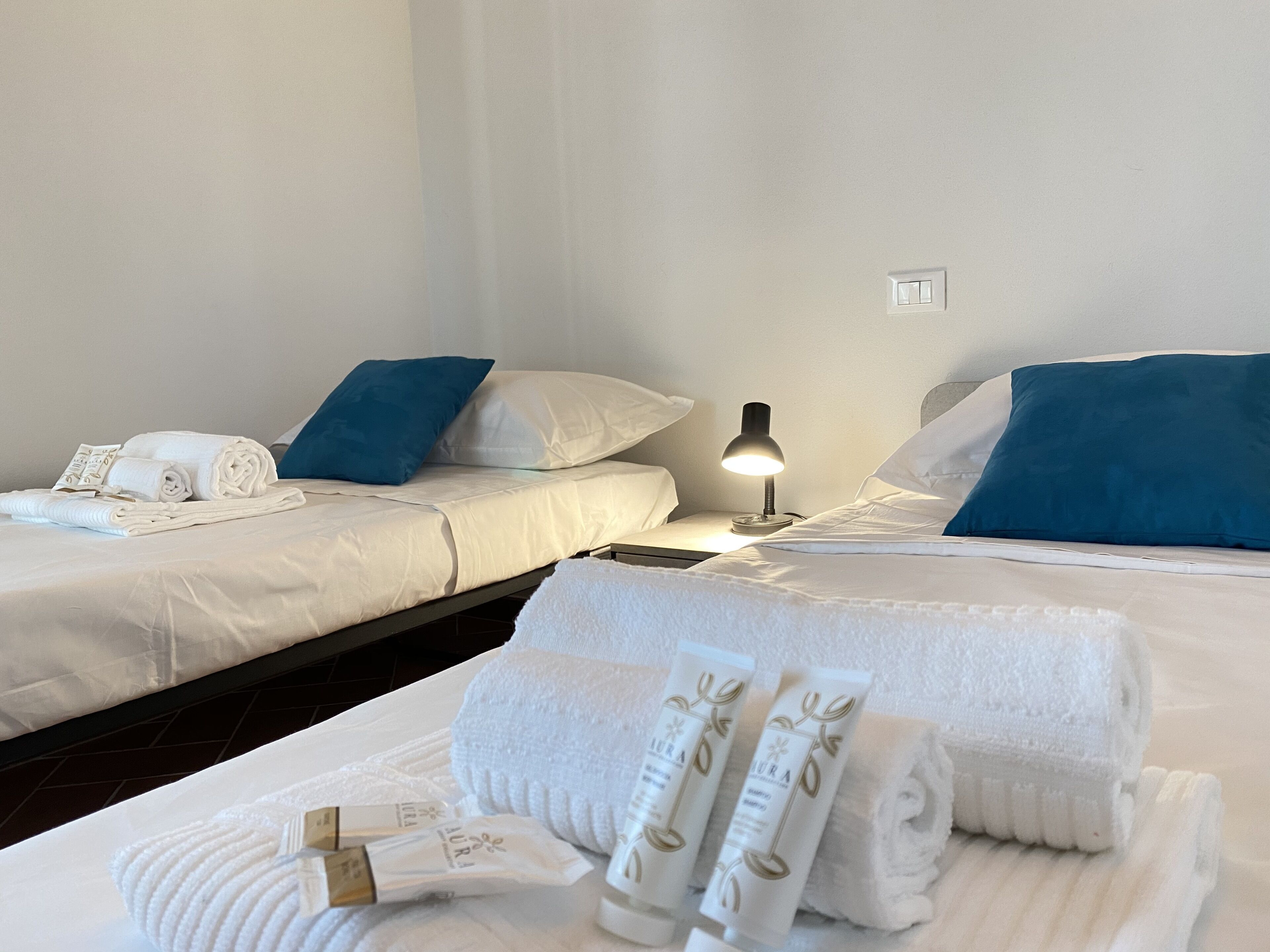 Ein Hotelzimmer mit zwei Betten, jedes mit weißen Laken und blauen Kissen. Ein Nachttisch mit Lampe, und Toilettenartikel sind ordentlich auf dem Bett angeordnet.