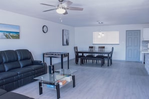 A modern living room with a black leather sofa, a glass coffee table, a dining area with a wooden table and chairs, a ceiling fan, and a kitchen area with a door and cabinets.