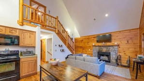 A spacious living area with a wooden staircase, a stone fireplace, a kitchen with modern appliances, and a cozy seating arrangement. - Creekside Cabin • Hot Tub • Decks (Maggie Valley)