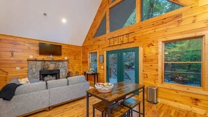 A cozy cabin interior with a stone fireplace, wooden walls, a gray sofa, and large windows offering a view of the forest. - Creekside Cabin • Hot Tub • Decks (Maggie Valley)