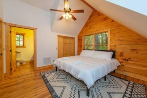 A bedroom with a sloped ceiling, wooden walls, a bed with white bedding, a ceiling fan, and a window with a view of trees.