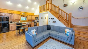 A spacious living area with a large gray sectional sofa, wooden flooring, a kitchen with wooden cabinets, a dining table, and a staircase with wooden railings. - Creekside Cabin • Hot Tub • Decks (Maggie Valley)