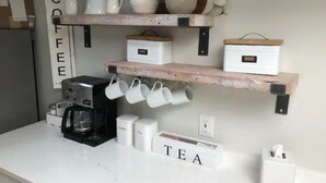 A kitchen with white countertops, a coffee maker, mugs hanging, and shelves with kitchenware.