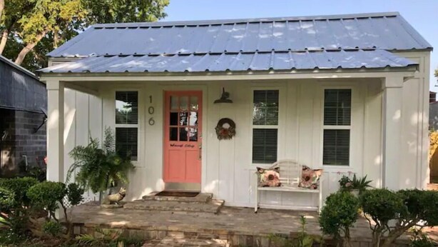 A white house with a red door, a porch with two chairs, and a number 106 on the wall.
