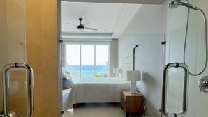 Un baño moderno con cabina de ducha de vidrio, un dormitorio visible con cama y vista al mar, y un ventilador de techo.