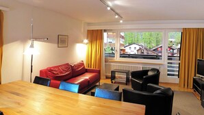 A room with a wooden dining table, red leather sofa, black chairs, a TV, and a window with a view of houses and trees. - Cristal by Interhome (Saas-Fee)