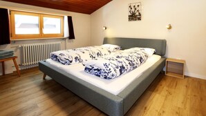 Une chambre avec un plafond en bois, un lit recouvert d’une literie à motifs floraux, une fenêtre avec des rideaux, un radiateur et une petite table de chevet.