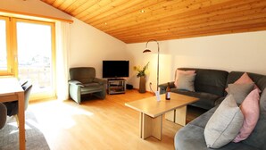 Un salon moderne avec un plafond en bois, un canapé gris, un meuble TV noir et un coin repas agrémenté d’un tapis à motifs étoilés.