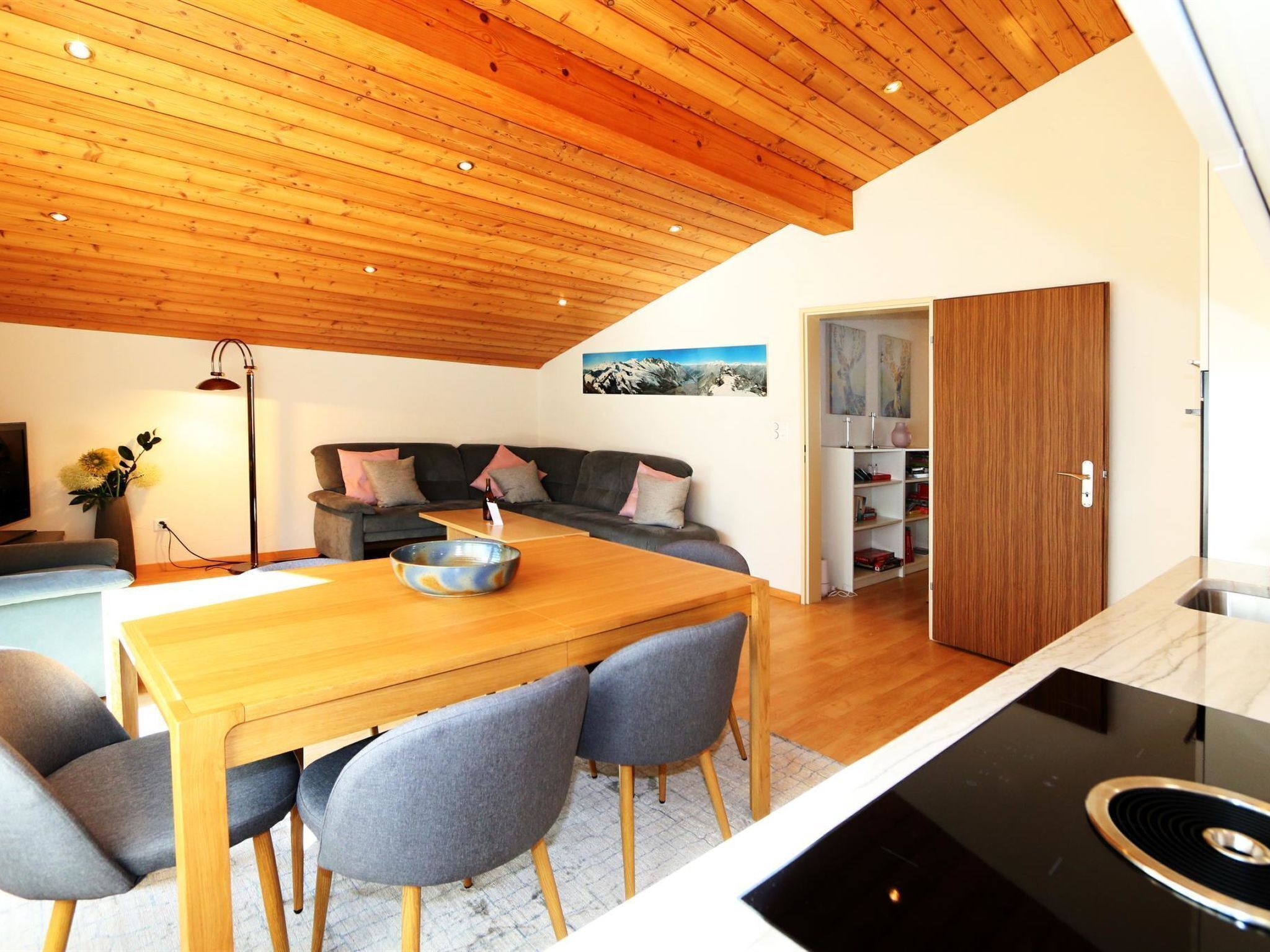 A modern kitchen with a wooden ceiling, a dining area with a table and chairs, and a living area with a sofa and a painting on the wall.