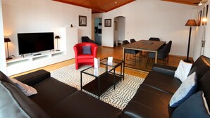 Ein modernes Wohnzimmer mit Flachbildfernseher, einem roten Sessel, einer Sofagarnitur, einem Esstisch mit Stühlen und einer Küche im Hintergrund.
