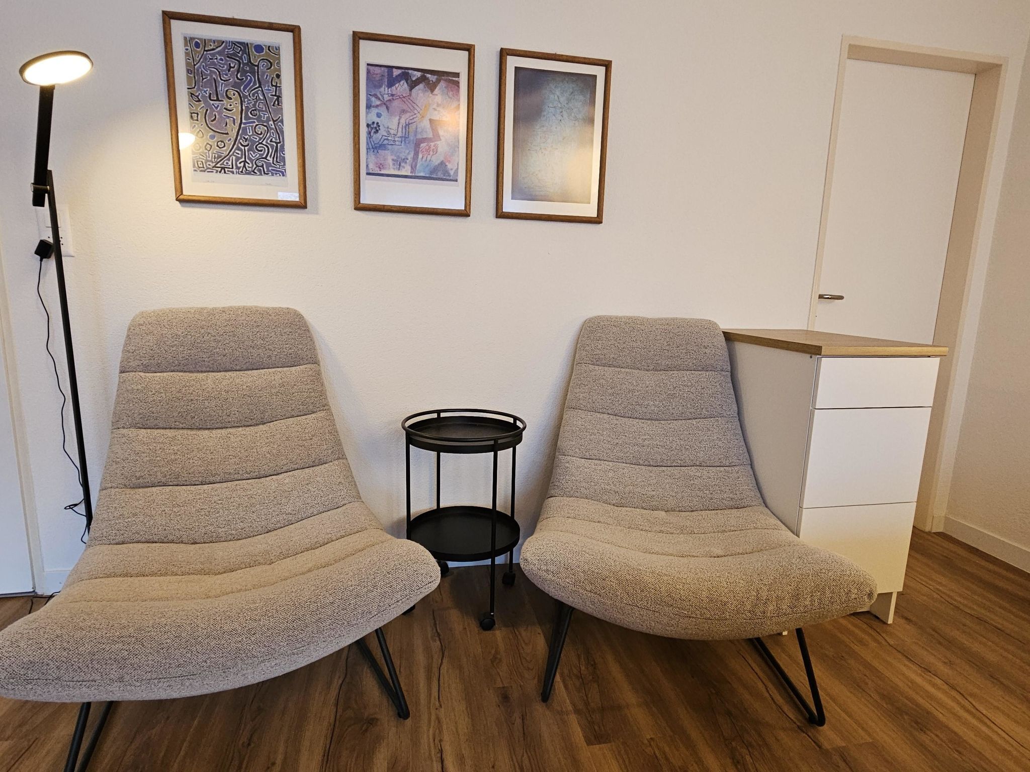 Dos sillones beiges con patas negras, una mesita auxiliar negra y tres obras de arte enmarcadas en la pared.