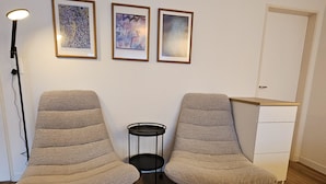 Dos sillones beiges con patas negras, una mesita auxiliar negra y tres obras de arte enmarcadas en la pared.