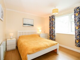 Un dormitorio con una cama blanca, una mesita de noche con una lámpara, una mesita auxiliar pequeña con una planta, una ventana con cortinas y un cuadro en la pared.