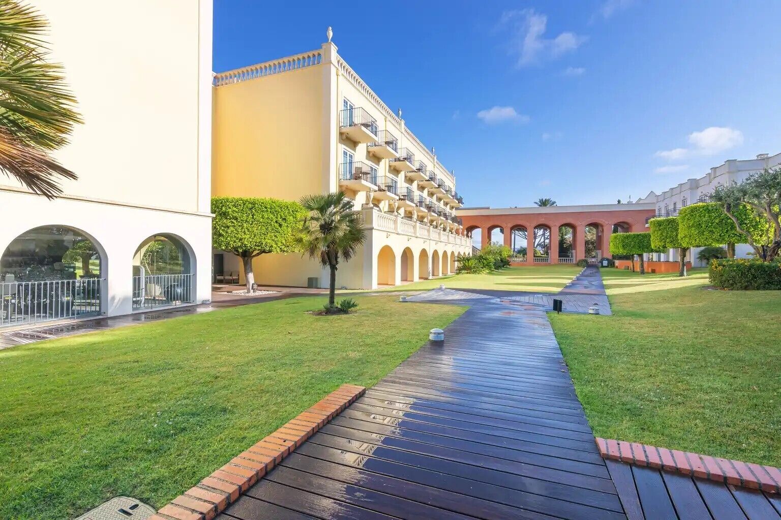A courtyard with a wooden pathway, palm trees, and a building with arched windows and balconies.