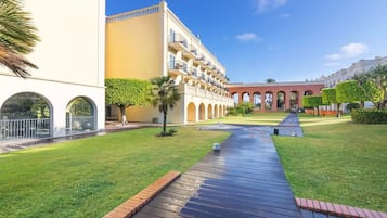 A courtyard with a wooden pathway, palm trees, and a building with arched windows and balconies.