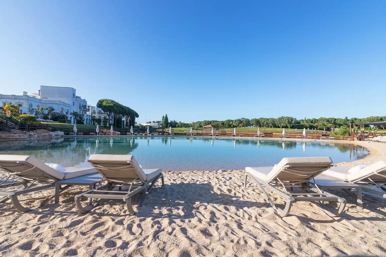 A poolside area with lounge chairs, a sandy beach, and a clear blue sky.