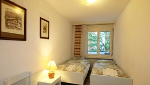 Ein kleiner, gut beleuchteter Raum mit zwei Einzelbetten, einem Nachttisch mit Lampe und einem Fenster mit Blick auf Bäume.