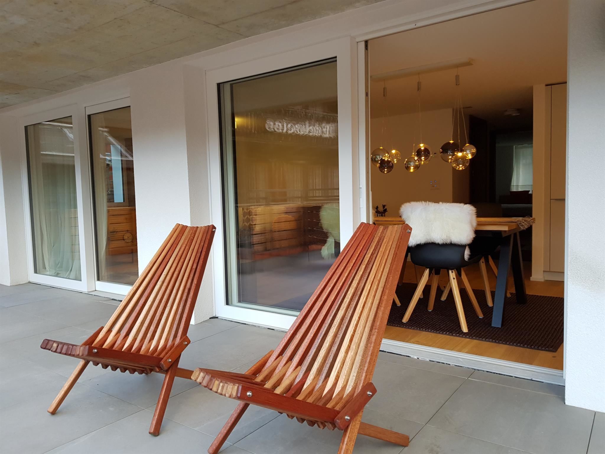 Un intérieur moderne avec deux fauteuils de salon en bois, un coin repas avec une chaise blanche à poils longs et une porte en verre donnant sur une pièce agrémentée d’un lustre.