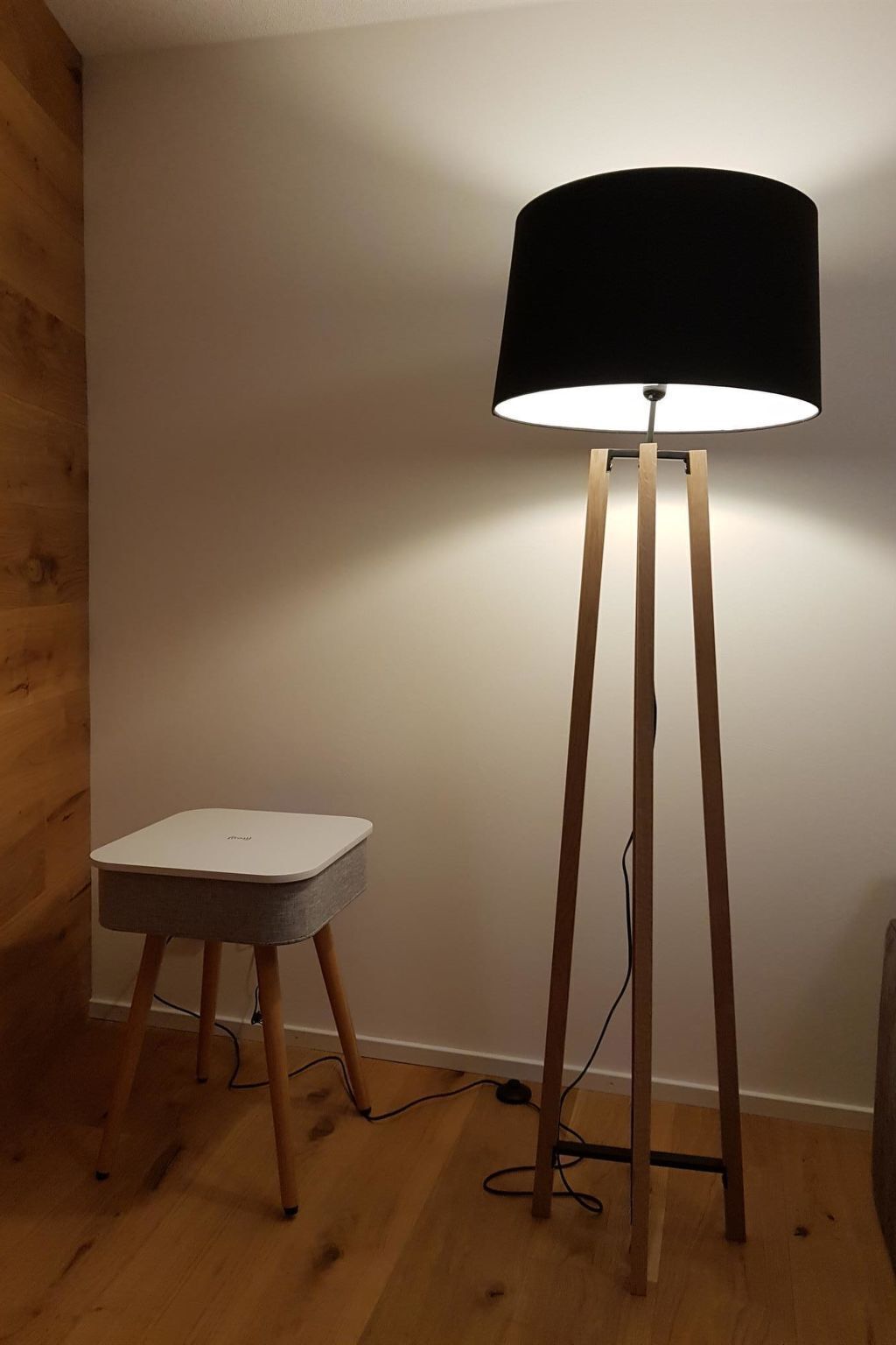 Eine Stehlampe mit schwarzem Lampenschirm und weißem Fuß, verbunden durch ein Kabel auf Holzboden.