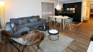 A modern living room with a grey sofa, a dining table with chairs, a small round table with a black base, and a pet dog. - Apartment Apollo by Interhome (Saas-Fee)