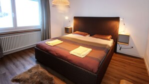 A modern bedroom with a large bed, a hanging light fixture, a nightstand, and a view of the outdoors through a window. - Apartment Apollo by Interhome (Saas-Fee)