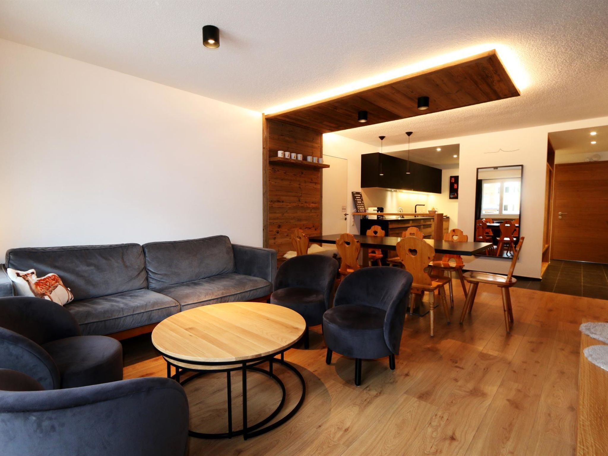 A modern living room with a grey sofa, round wooden coffee table, and dark armchairs. A kitchen with wooden cabinets and a dining area with wooden chairs is visible in the background.