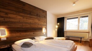 Cama de casal com roupa branca, cabeceira de madeira, luminárias de cabeceira, um banco e uma janela com cortinas.