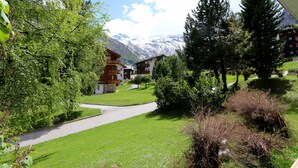 Uma pousada de montanha com um caminho pavimentado até os edifícios, cercada por vegetação exuberante e um cenário de picos nevados.