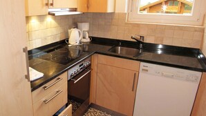 Een kleine keuken met een fornuis, spoelbak en vaatwasser, houten keukenkastjes en een raam met uitzicht op een huis.