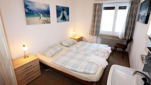 Een slaapkamer met een bed, nachtkastjes, een lamp, een raam met gordijnen, een stoel, en een badkamer met een wastafel en spiegel.