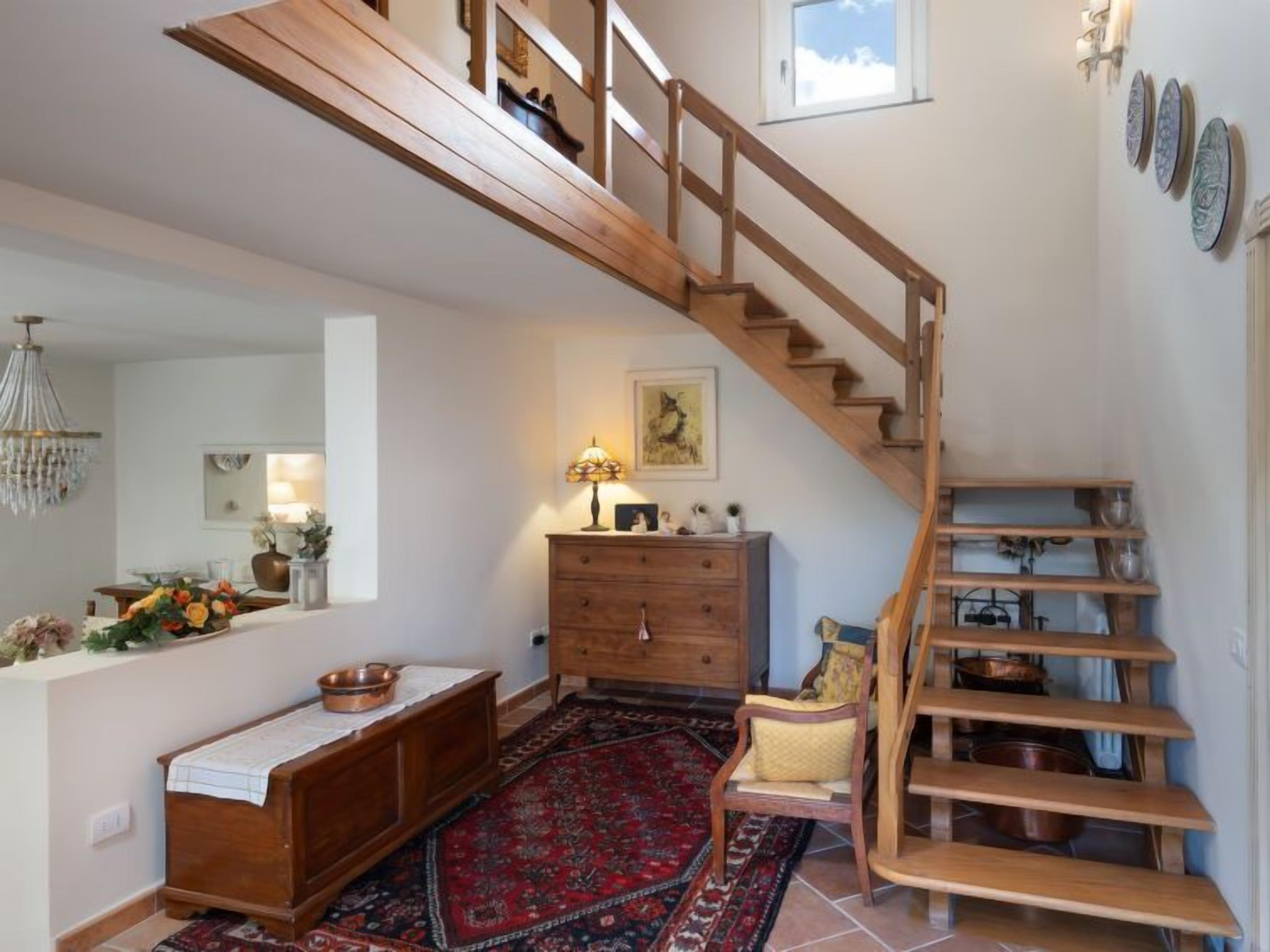 Eine Treppe mit Holzwangen führt zu einer oberen Etage, eine Holzkommode mit Schubladen, ein kleiner Tisch mit einer Lampe und ein Teppich mit Muster auf dem Boden.