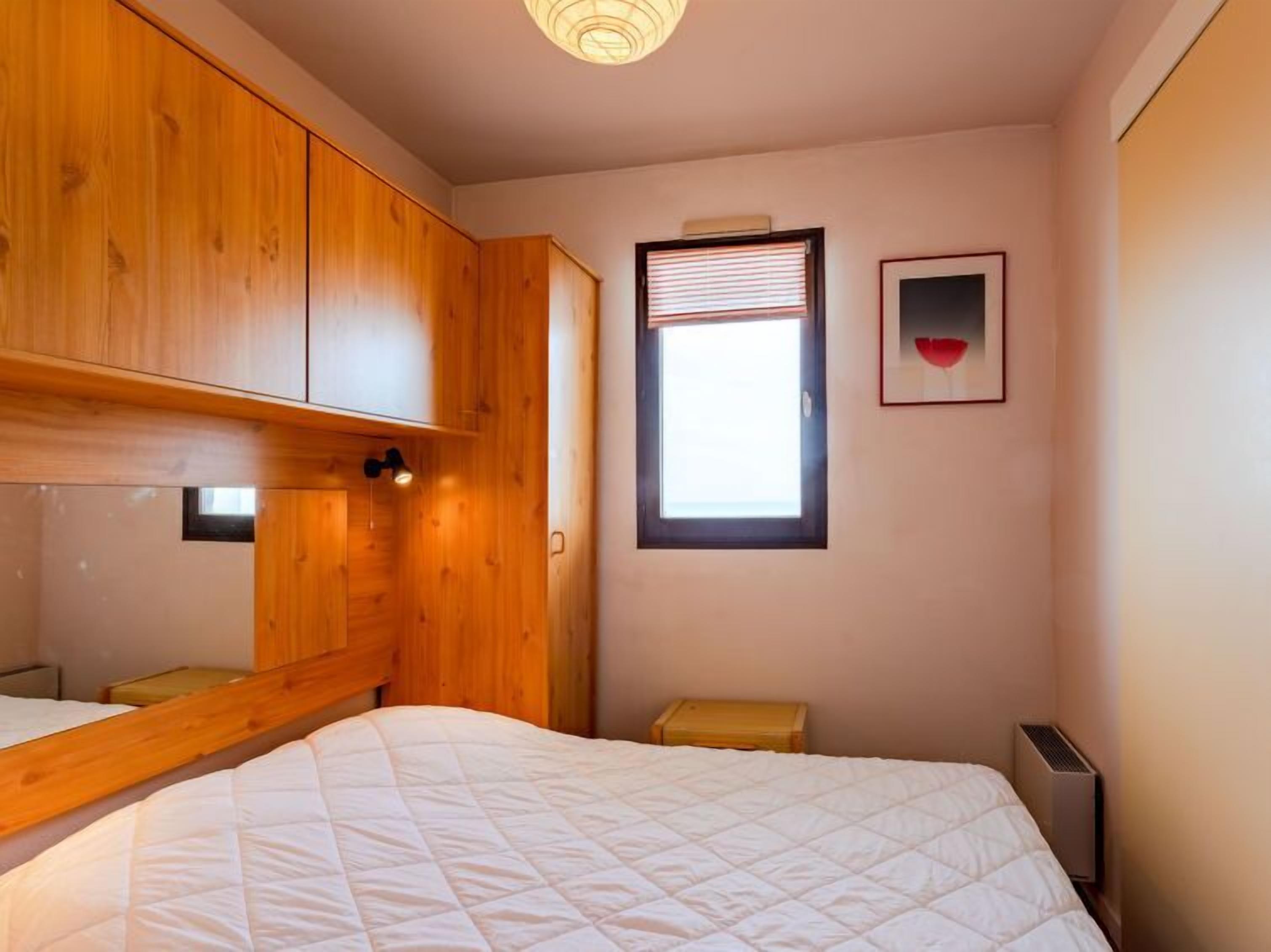 A bedroom with a wooden wardrobe, a bed with a white quilt, a window with blinds, and a radiator.