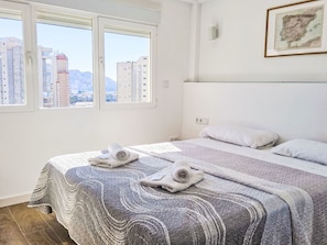 Ein Schlafzimmer mit einem Bett, zwei Kissen, einer Decke und einem Handtuch. Zwei Fenster bieten Blick auf die Stadt.