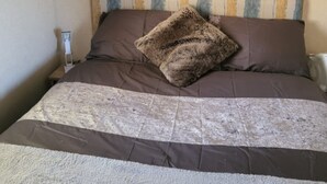 Una cama con cabecera de madera, una colcha marrón y beige, y una sola almohada marrón. Encima de la cama hay un estante con dos velas y un pequeño florero.