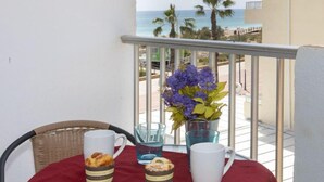 A balcony with a table set for breakfast, featuring a wicker chair, a red tablecloth, two mugs, a glass of water, and a small basket of pastries. - Abanicos by Interhome (Peñiscola)