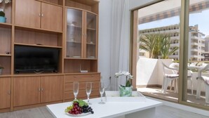 A modern living room with a flat-screen TV, wooden cabinets, a white table with a fruit platter and glasses, and a balcony with outdoor furniture. - Peñismar I by Interhome (Peñiscola)