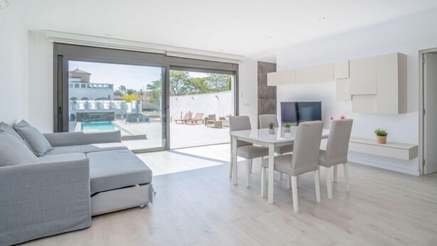 Un salon moderne avec un canapé gris, une table à manger et des chaises, un téléviseur à écran plat fixé au mur et une porte-fenêtre coulissante donnant sur une zone extérieure avec piscine.