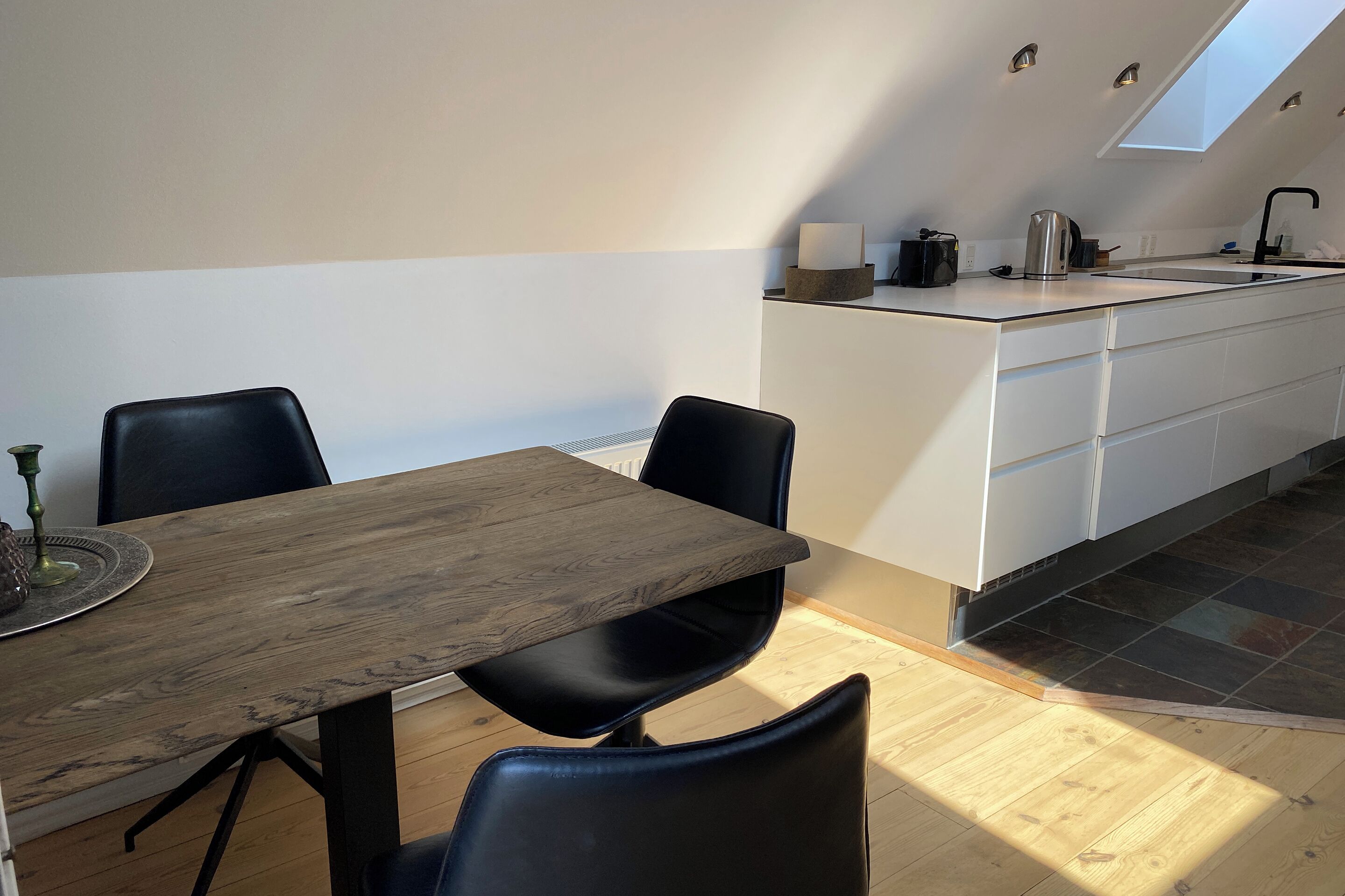 Una cocina moderna con tragaluz, un comedor con mesa de madera y sillas negras, y una pequeña isla con un hervidor.