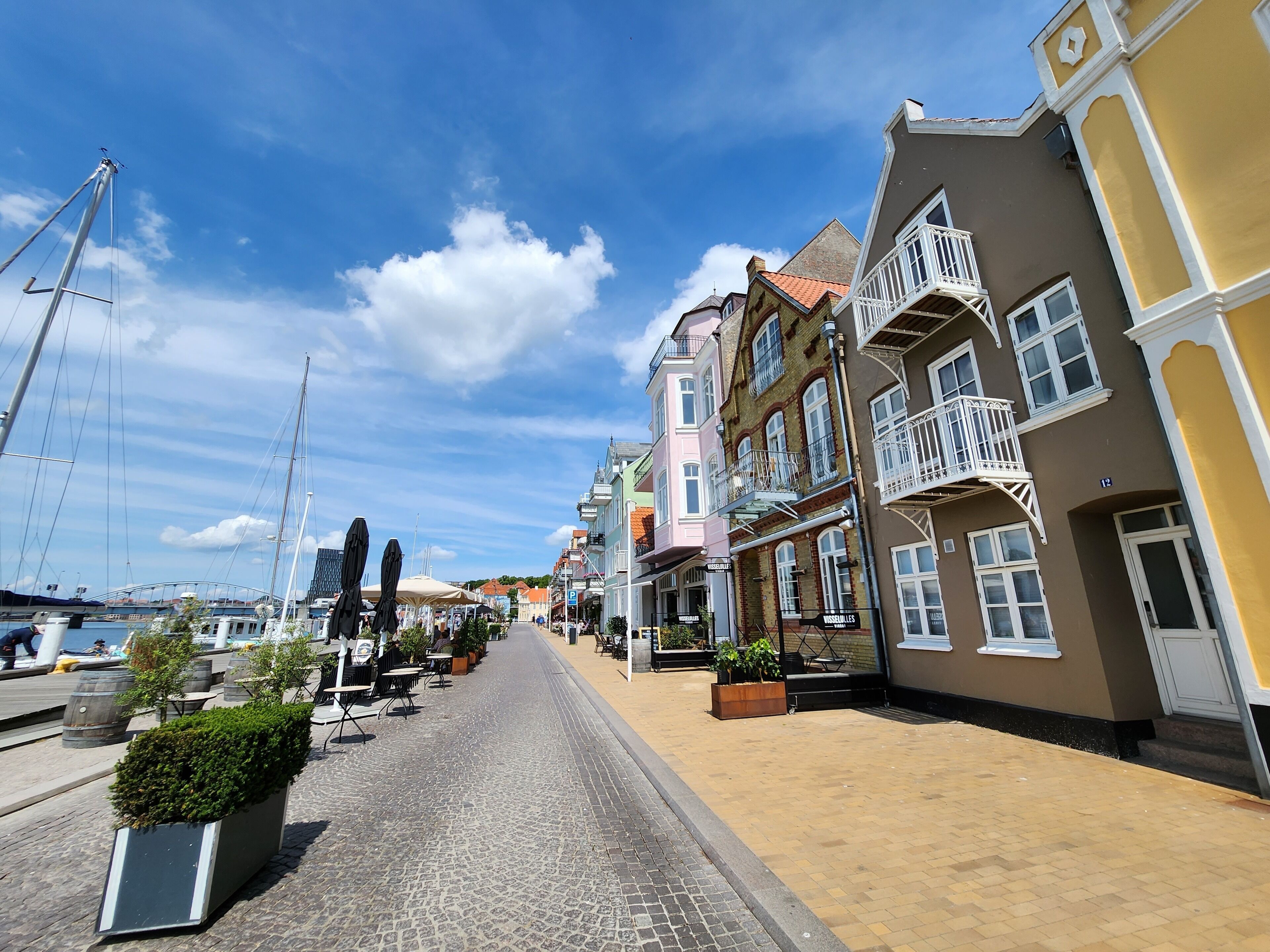A cobblestone street lined with colorful buildings, some with balconies and outdoor seating areas.