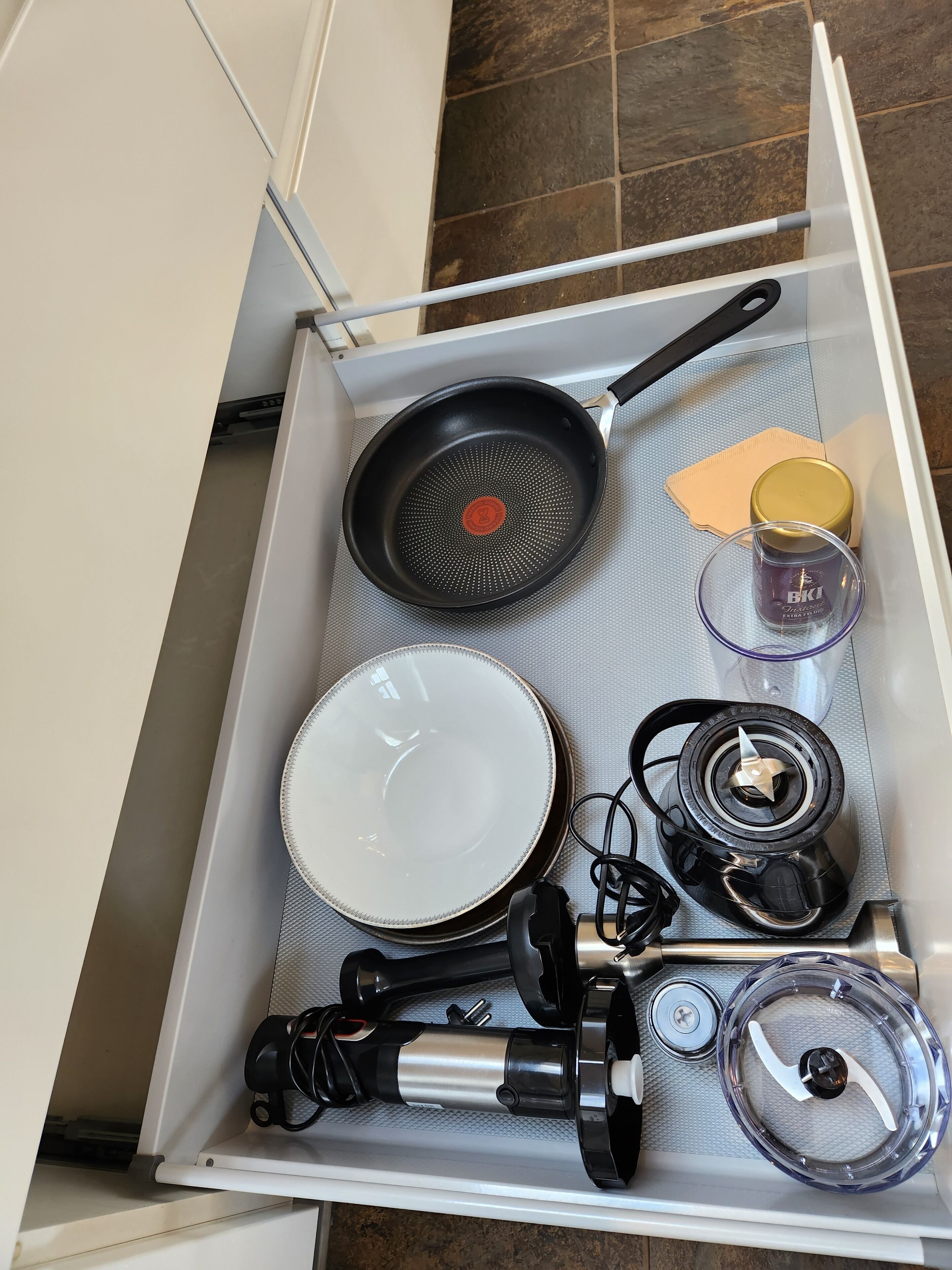 A kitchen drawer with a frying pan, a white plate, a blender, a canister, and a small container.