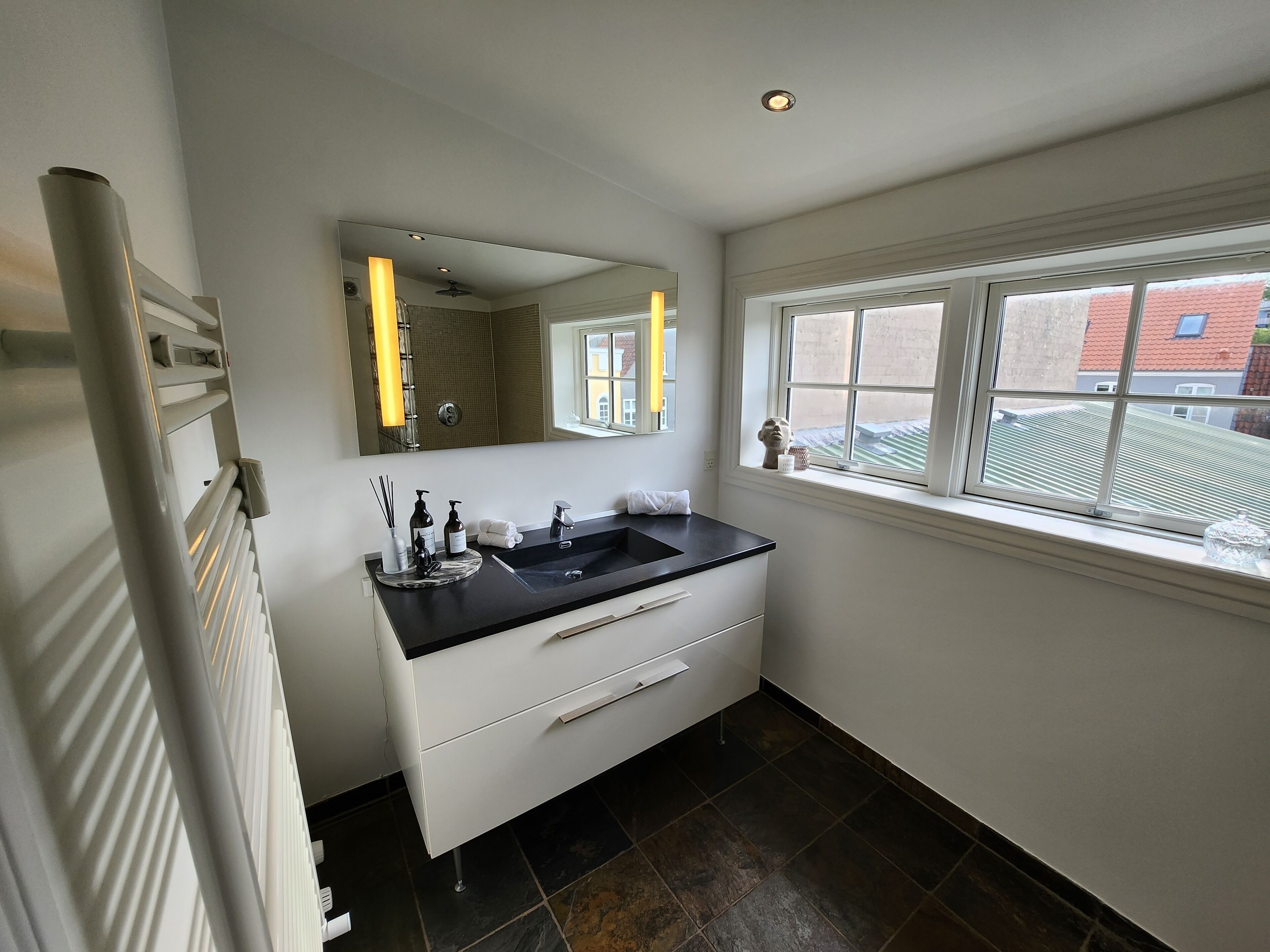 Een moderne badkamer met een zwart wastafelmeubel, witte kasten, een grote spiegel en een raam met uitzicht op gebouwen.