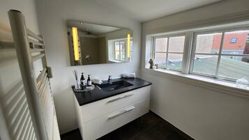 Een moderne badkamer met een zwart wastafelmeubel, witte kasten, een grote spiegel en een raam met uitzicht op gebouwen.
