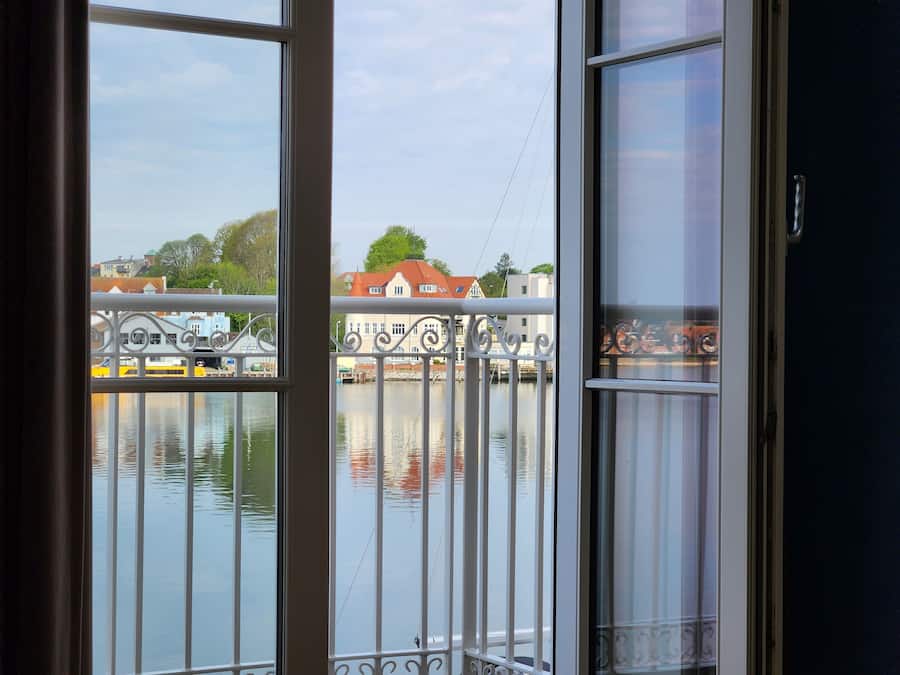 A view from a window with white railings overlooking a calm body of water, with buildings and trees in the background.